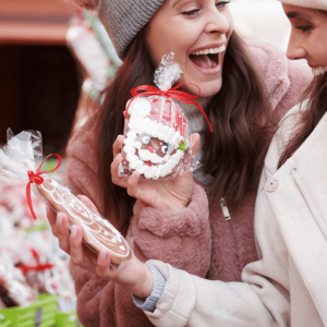 Two women smiling and holding festive holiday treats, including a gingerbread cookie and a Santa-shaped candy, at a Christmas market near the NJ Shore.